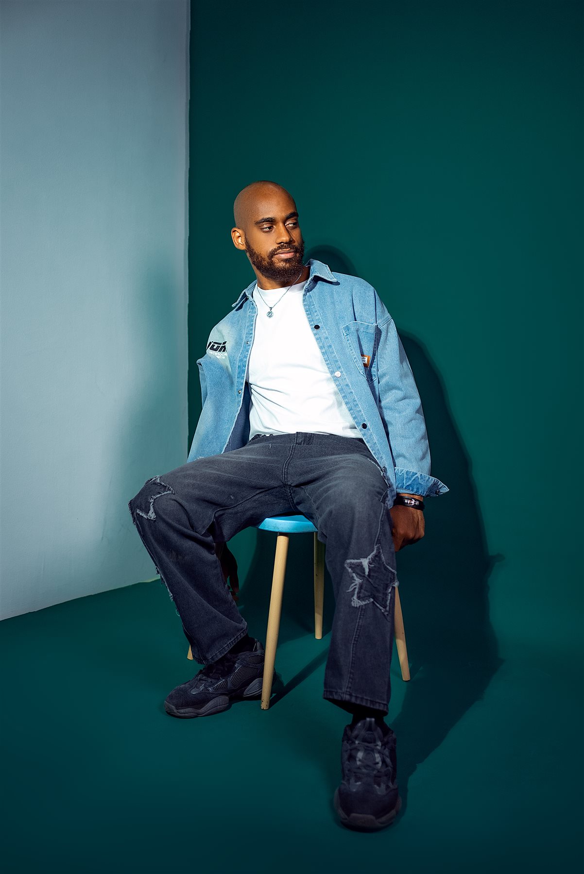 Moceol seated on a stool in a denim shacket, teal studio backdrop